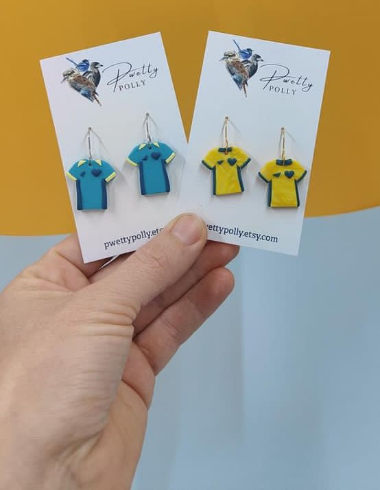 Teal and yellow Matilda shirt earrings on a hand with a yellow background