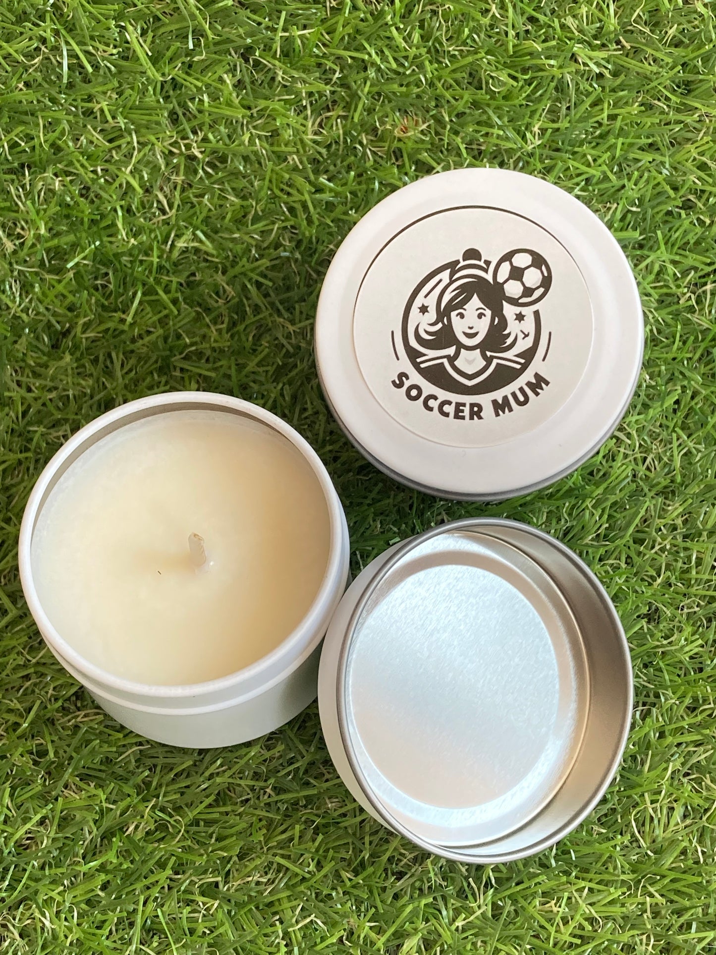 Candle in a white tin with a Soccer Mum logo on top laying on the grass.