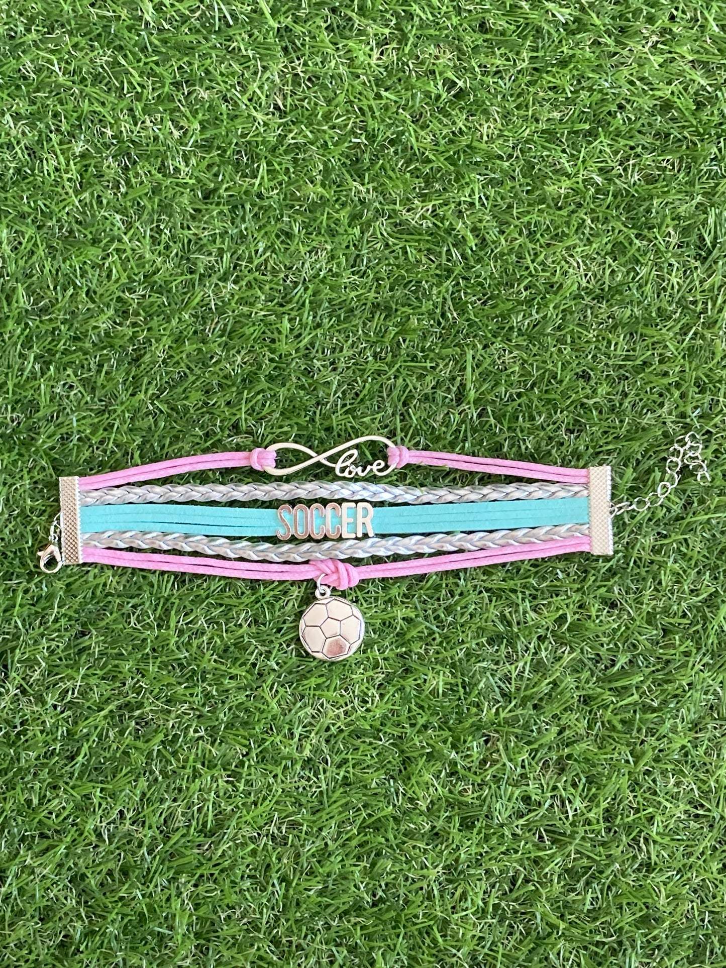 Up close image of pink and teal  bracelet with 3 soccer charms saying, soccer, love and a soccer ball charms laying on soccer grass.