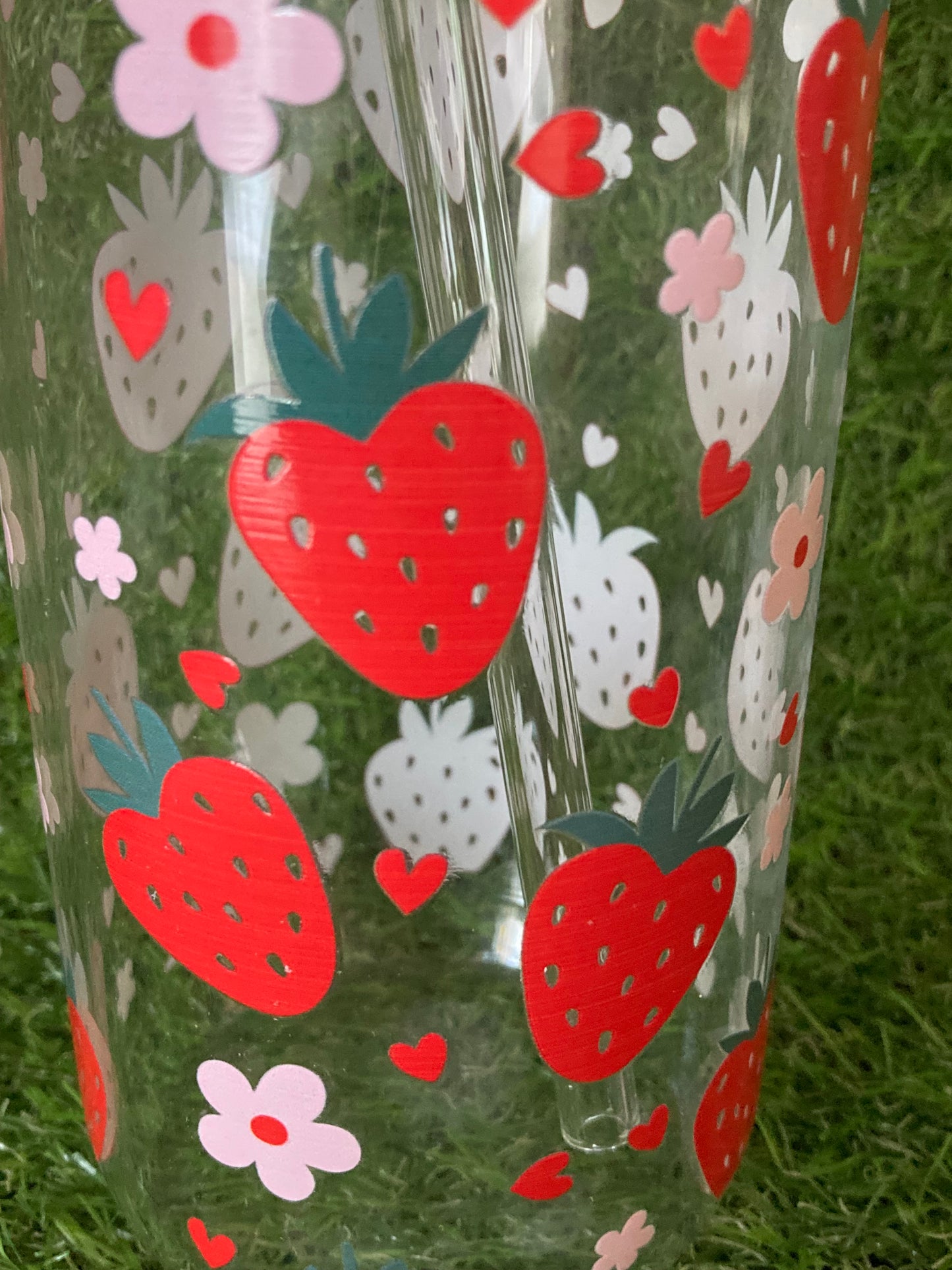 Close up of Clear glass can tumbler featuring strawberry and pink flower print with bamboo lid, clear straw and straw cleaner on green grass background.