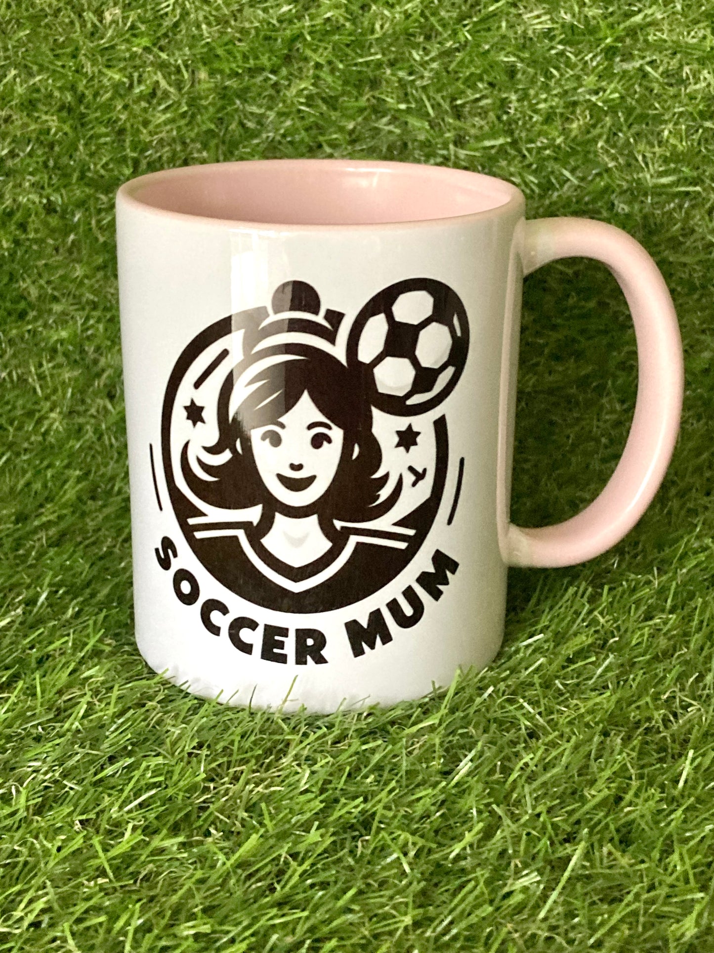 White mug with pink handle and insides with soccer mum logo printed on the side while sitting on grass.