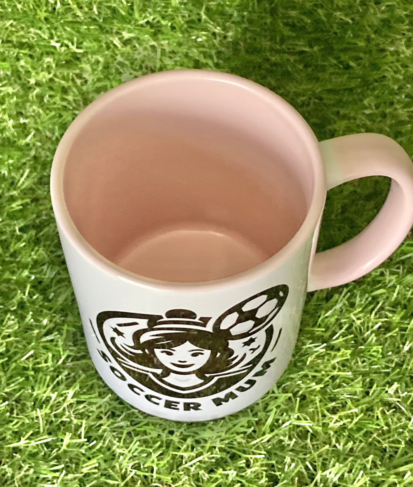 Top view of White mug with pink handle and insides with soccer mum logo printed on the side while sitting on grass.