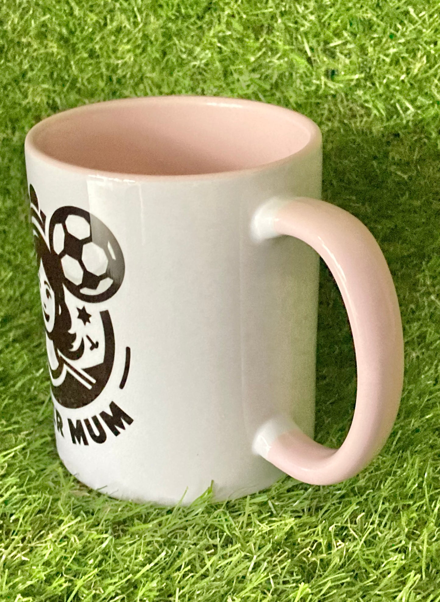 Side view of White mug with pink handle and insides with soccer mum logo printed on the side while sitting on grass.