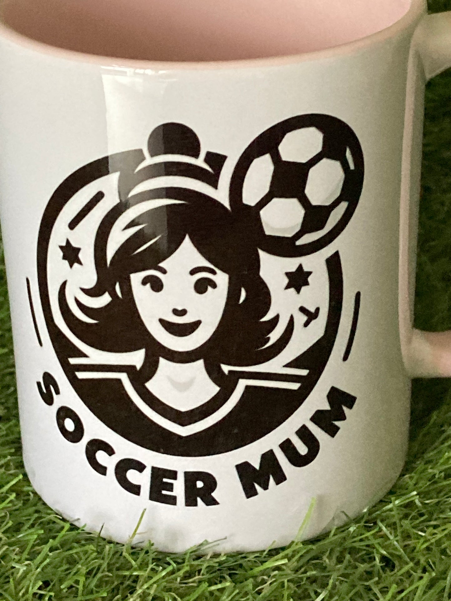 Close up of White mug with pink handle and insides with soccer mum logo printed on the side while sitting on grass.