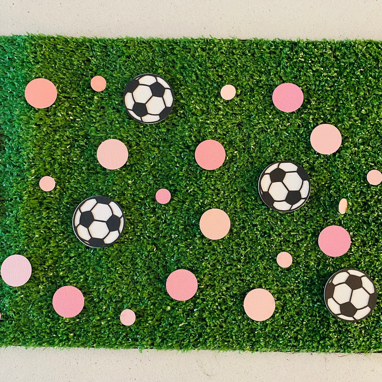 Large and small pink paper round discs with soccer ball paper discs scattered across green grass.