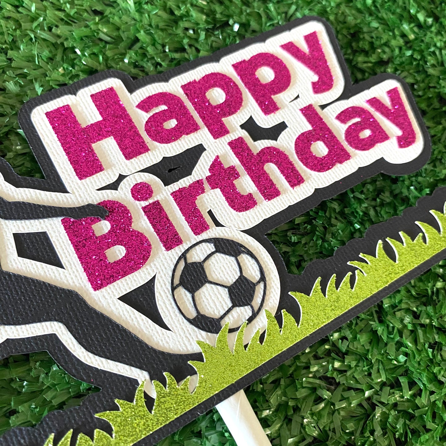 Female cake topper with pink words saying happy birthday and a girl soccer player in black silhouette kicking a soccer ball on glitter green grass.