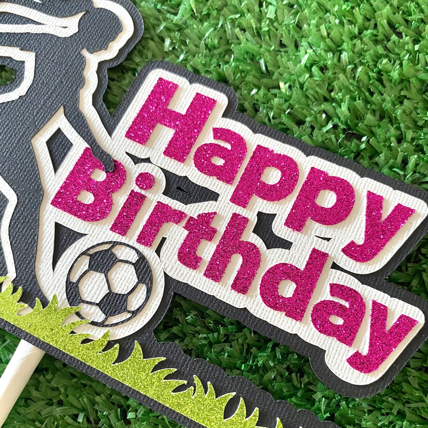 Female cake topper with pink words saying happy birthday and a girl soccer player in black silhouette kicking a soccer ball on glitter green grass.