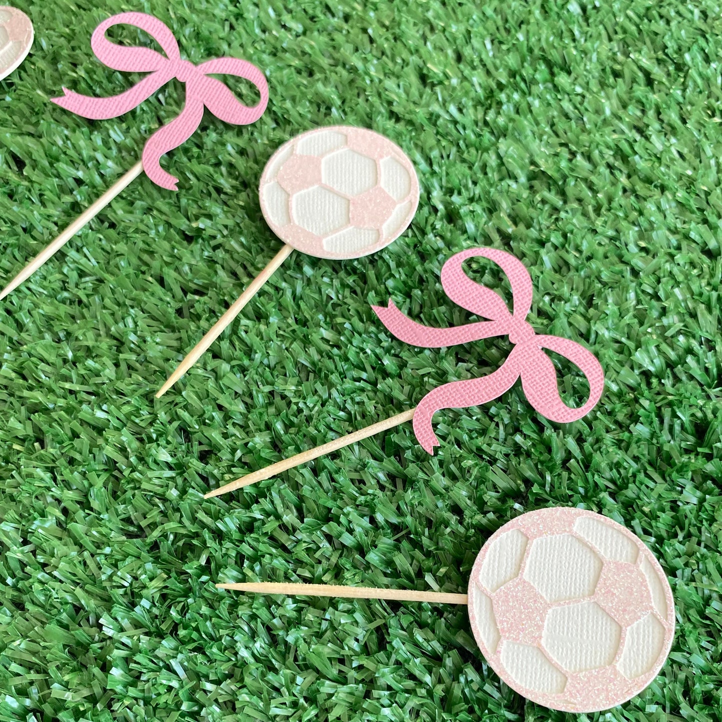 Pink and white Glitter soccer ball cup cake toppers with pink bow cup cake toppers laying out on top of green grass.