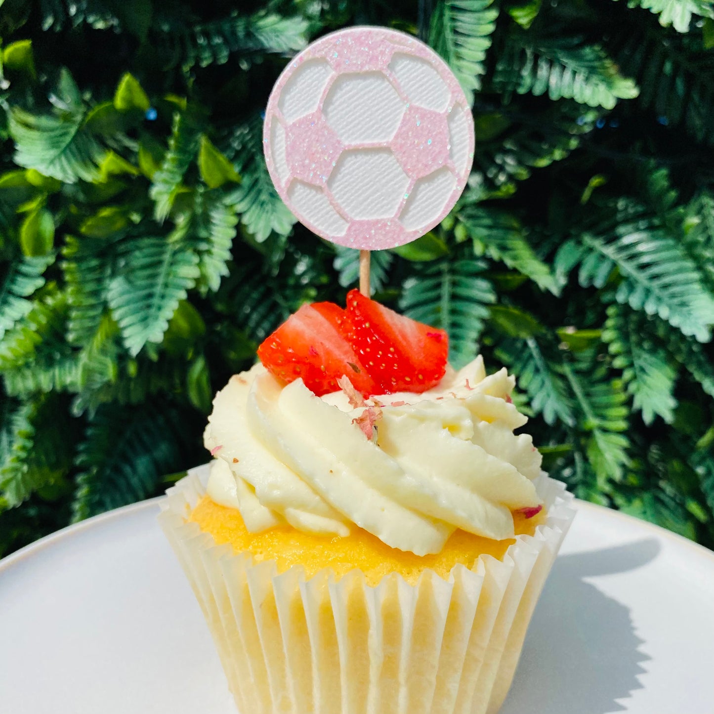 Pink and white Glitter soccer ball cup cake topper on top of a vanilla cupcakes with strawberry's on top in front on a green leaf back drop.