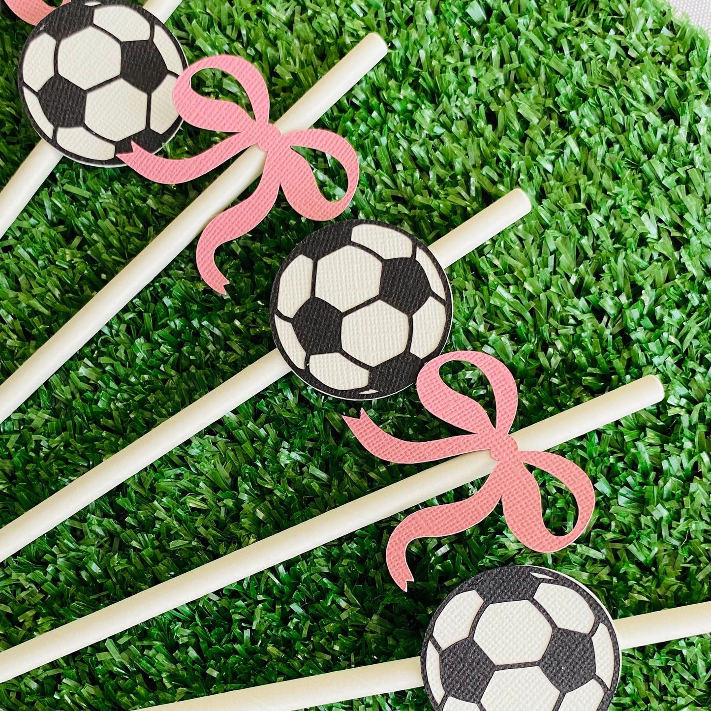 White straws topped with black and white soccer ball paper straw toppers and pink bow paper straw toppers laying on green grass in a fan shape.
