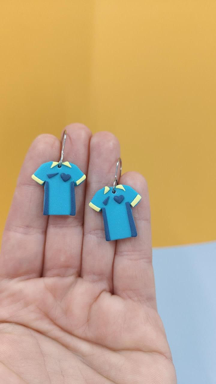 Teal and yellow Matilda shirt earrings on a hand with a yellow background