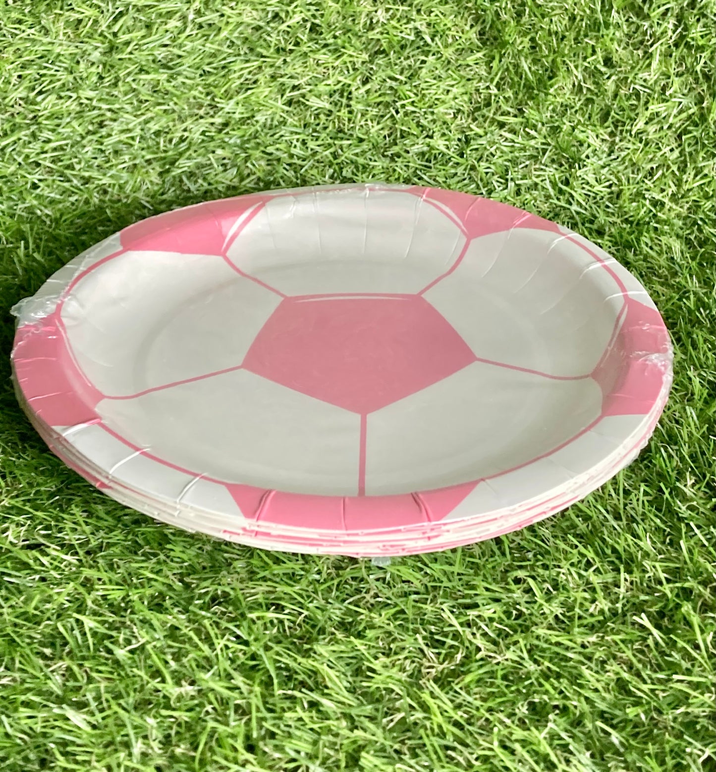 Pink soccer ball theme paper cake plate displayed on astro turf green grass. Side view.