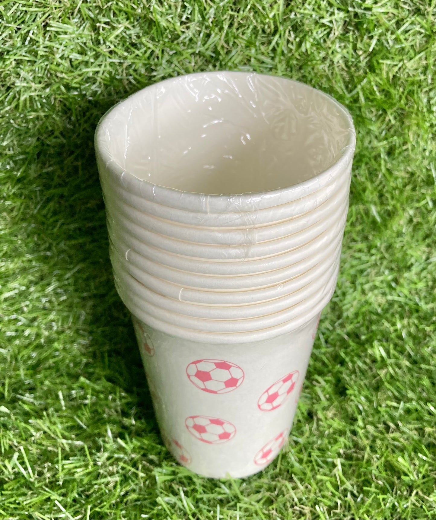 Pink soccer ball theme paper cups displayed on astro turf green grass. View from the top.