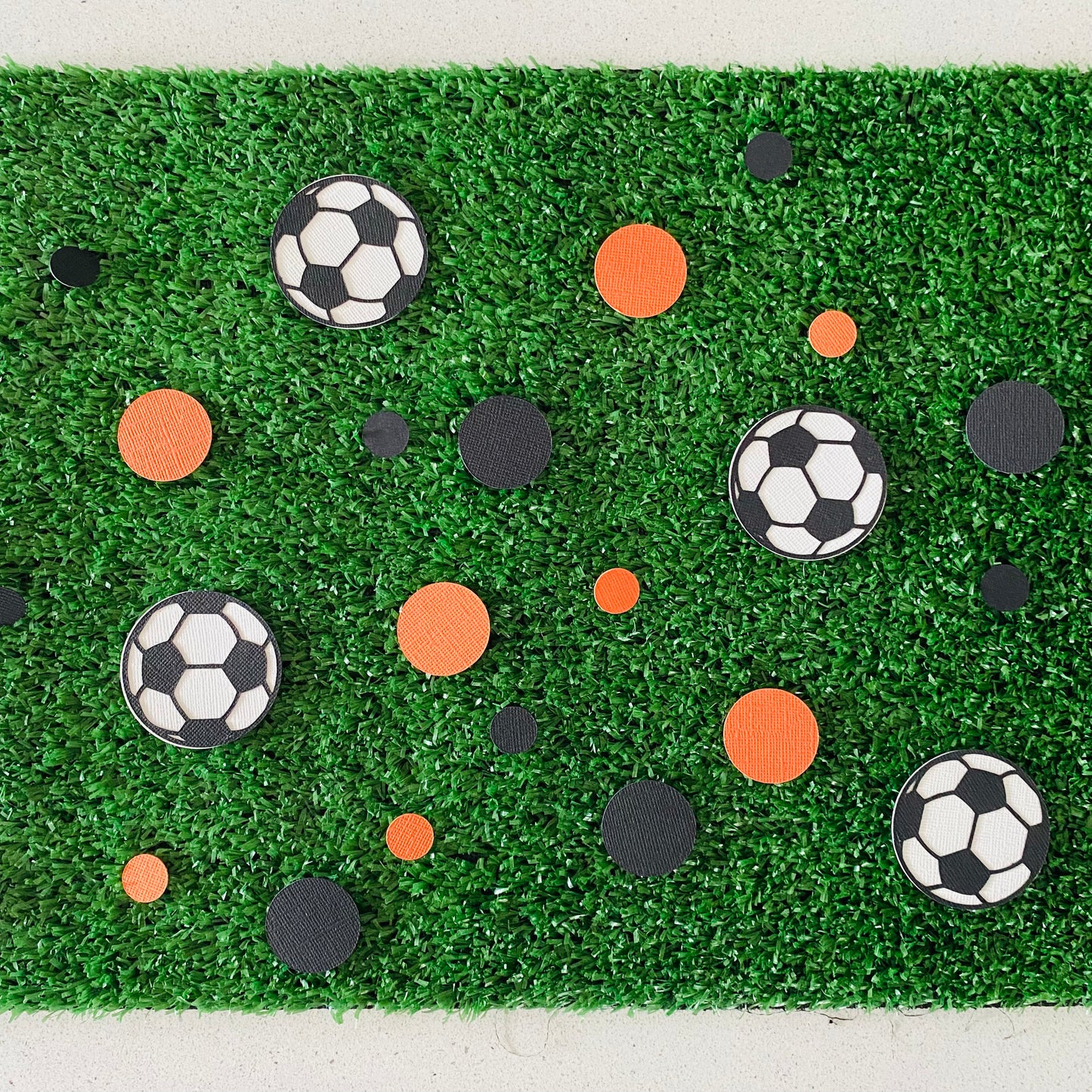 Orange and black paper round discs with soccer ball paper discs  scattered across green grass.