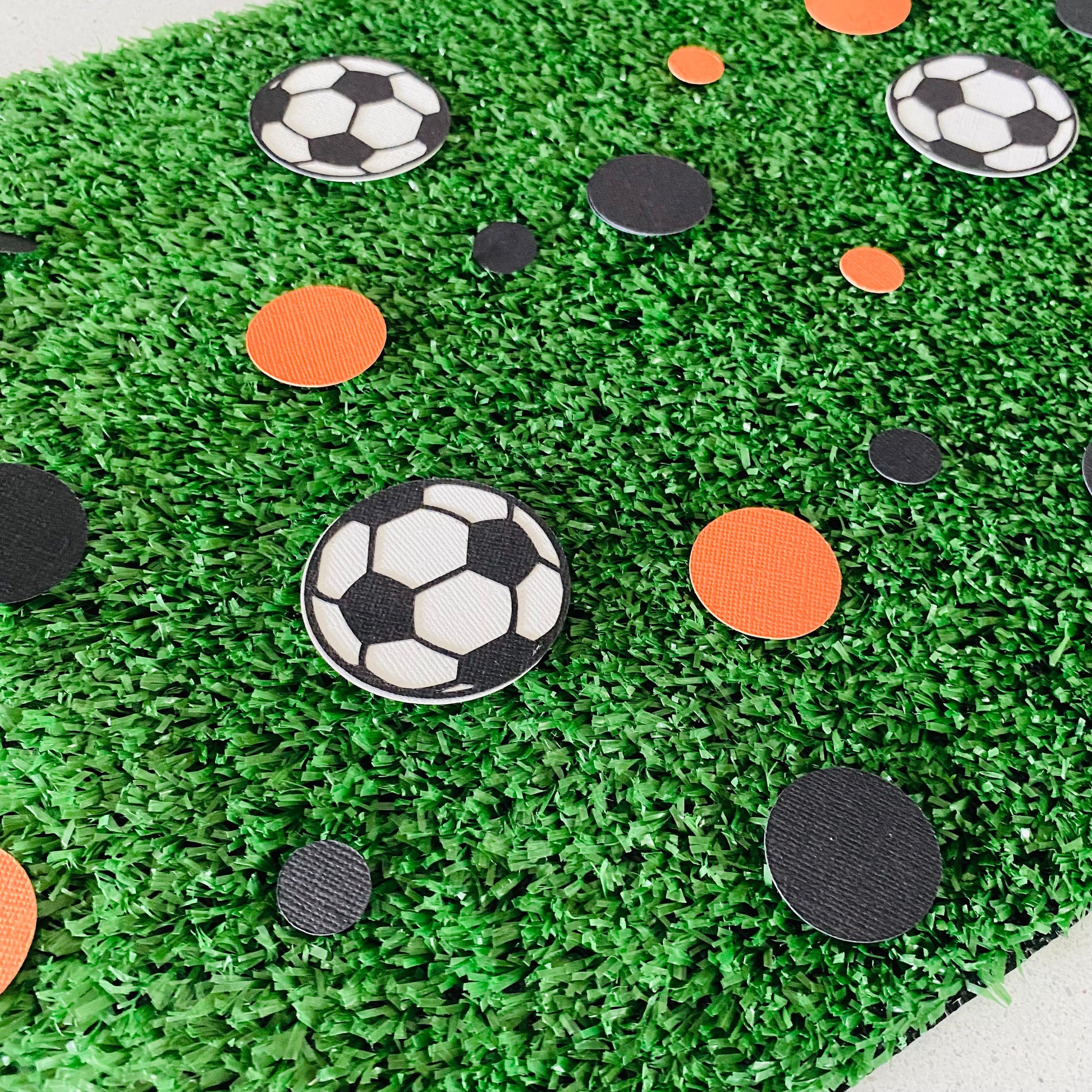 Orange and black paper round discs with soccer ball paper discs  scattered across green grass.