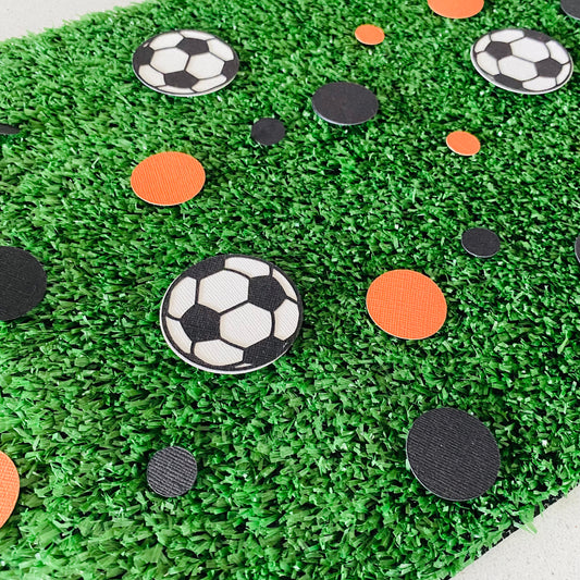 Orange and black paper round discs with soccer ball paper discs  scattered across green grass.
