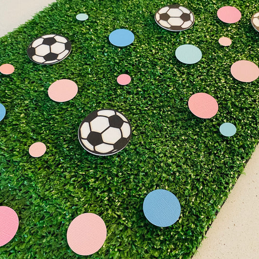 Large and small pink and blue paper round discs with soccer ball paper discs  scattered across green grass.
