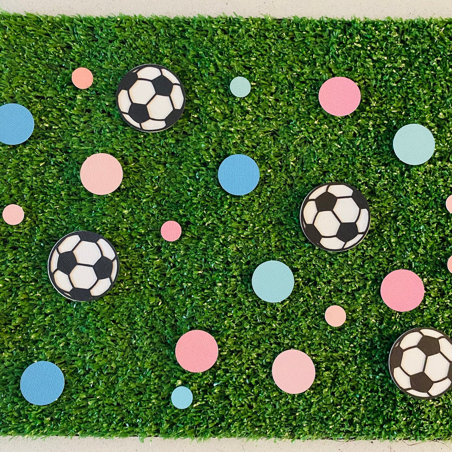 Large and small pink and blue paper round discs with soccer ball paper discs  scattered across green grass.