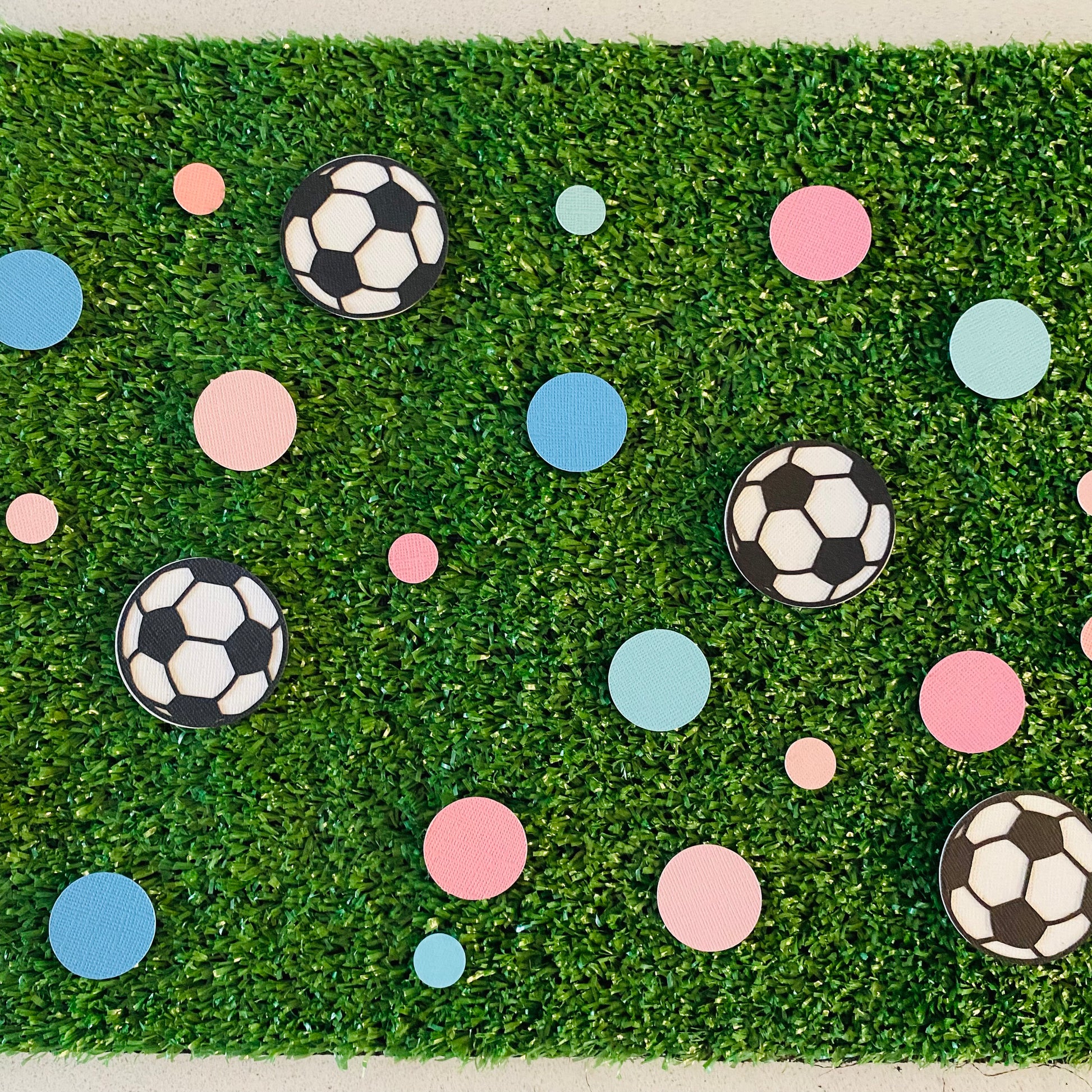 Large and small pink and blue paper round discs with soccer ball paper discs  scattered across green grass.