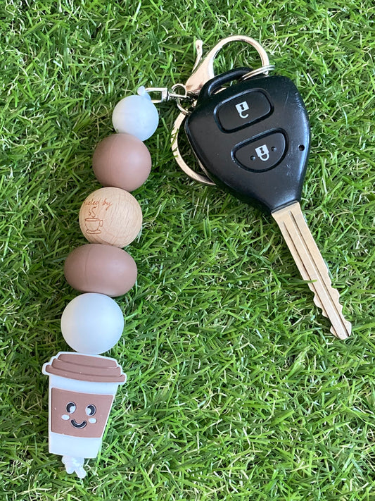 Brown and white beaded keychain with rubber coffee cup with smiley face with a car key laying on the grass.