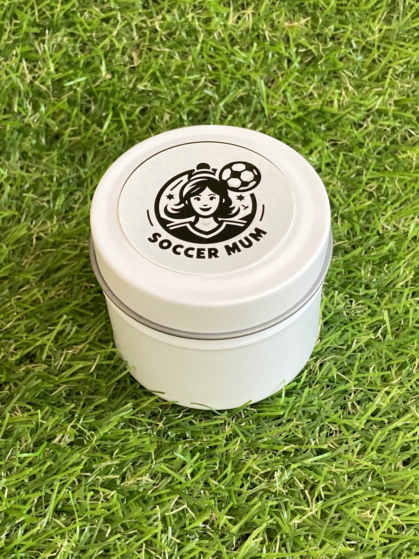 Candle in a white tin with a Soccer Mum logo on top laying on the grass.