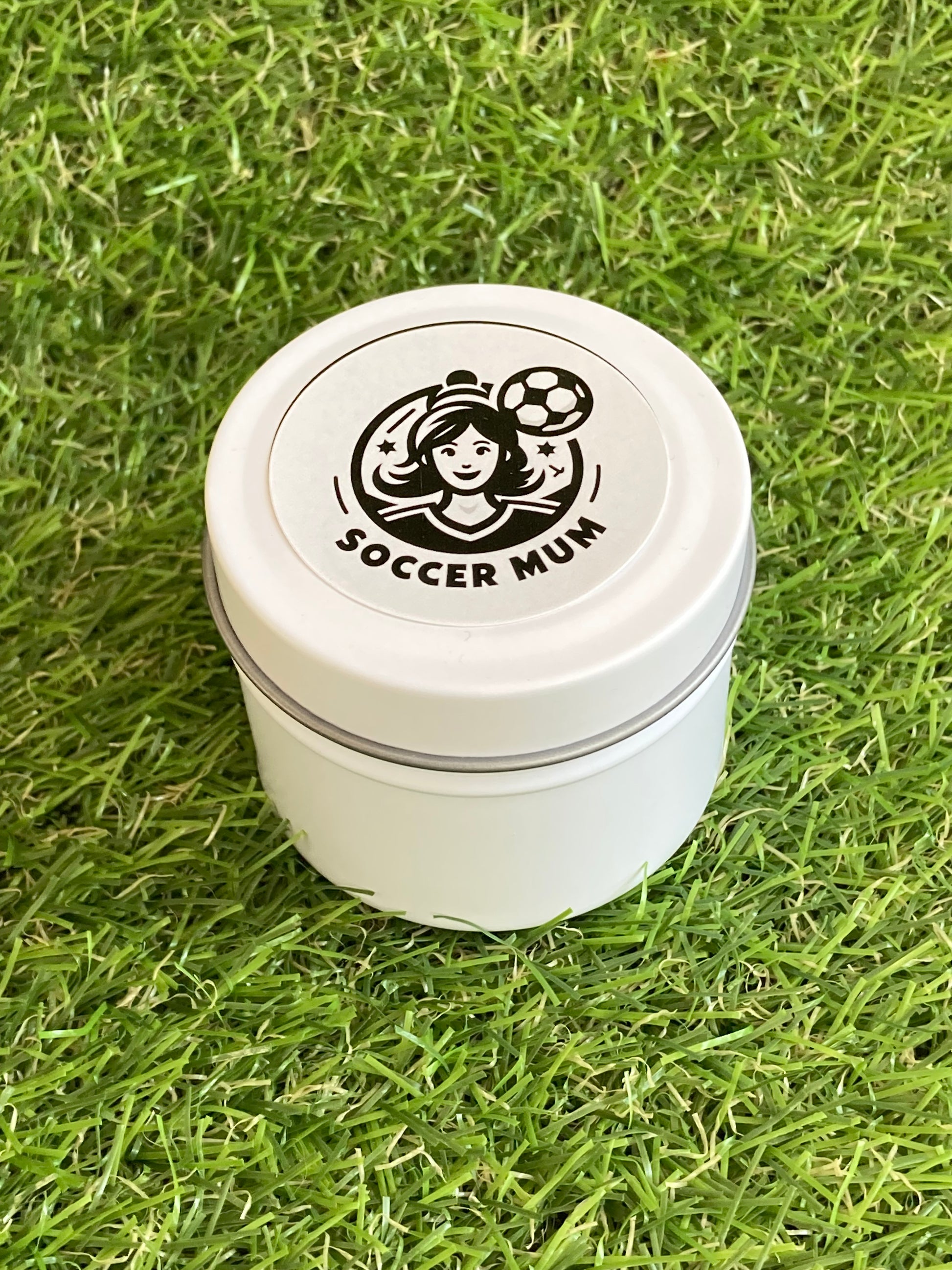 Candle in a white tin with a Soccer Mum logo on top laying on the grass.