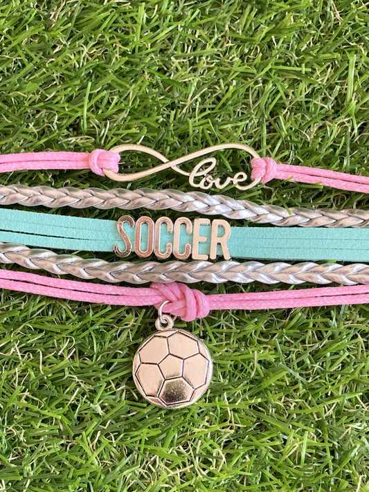 Up close image of pink and teal  bracelet with 3 soccer charms saying, soccer, love and a soccer ball charms laying on soccer grass.