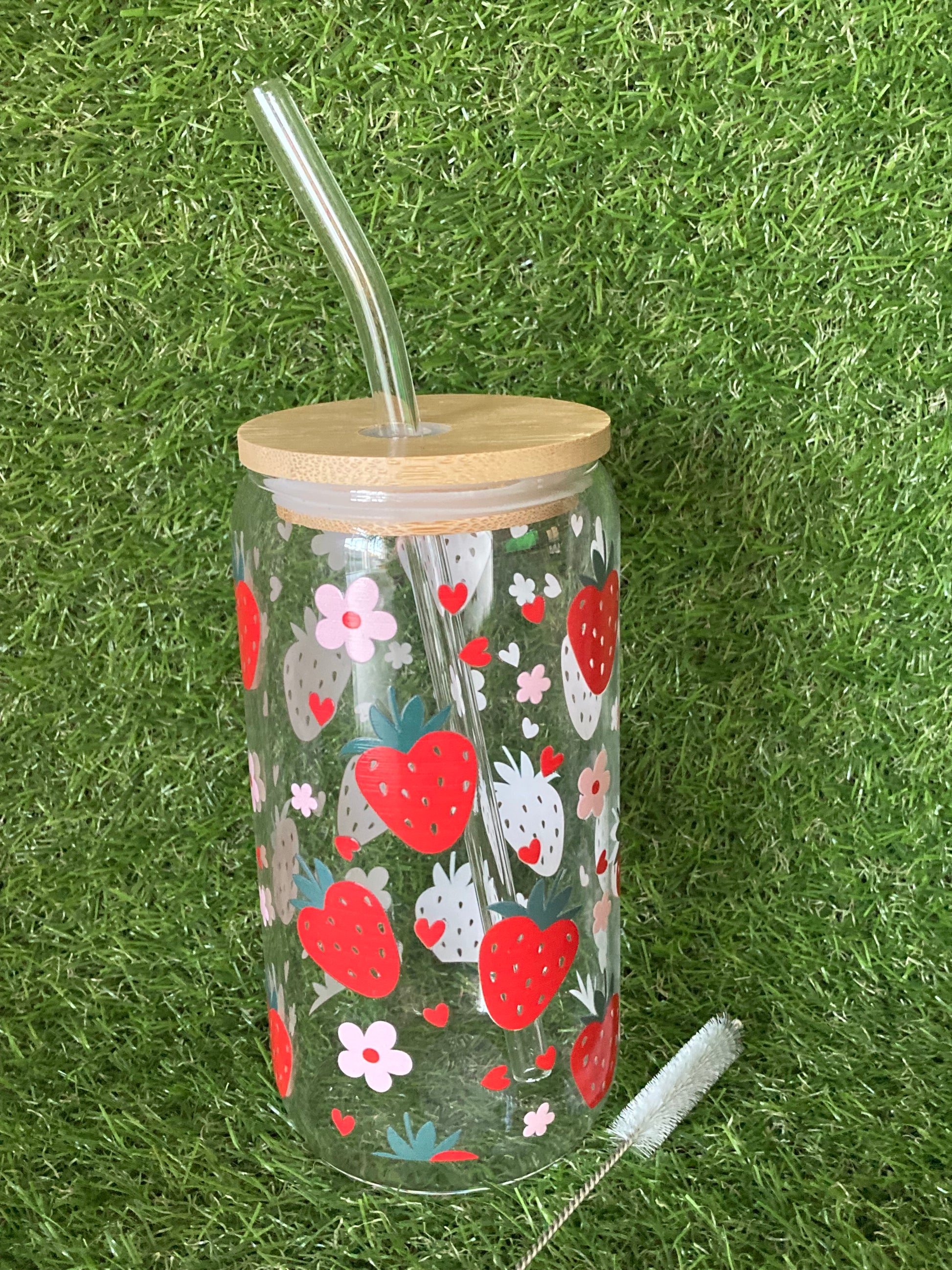 Clear glass can tumbler featuring strawberry and pink flower print with bamboo lid, clear straw and straw cleaner on green grass background.