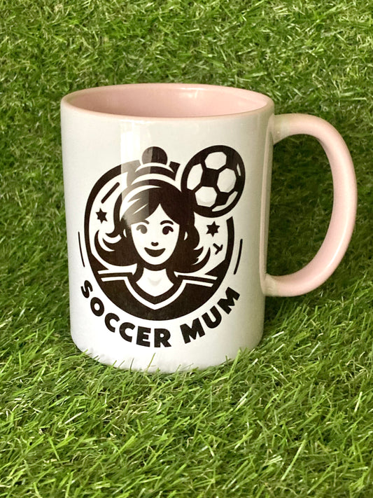 White mug with pink handle and insides with soccer mum logo printed on the side while sitting on grass.