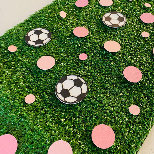 Large and small pink paper round discs with soccer ball paper discs  scattered across green grass.