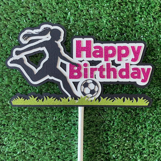 Female cake topper with pink words saying happy birthday and a girl soccer player in black silhouette kicking a soccer ball on glitter green grass.