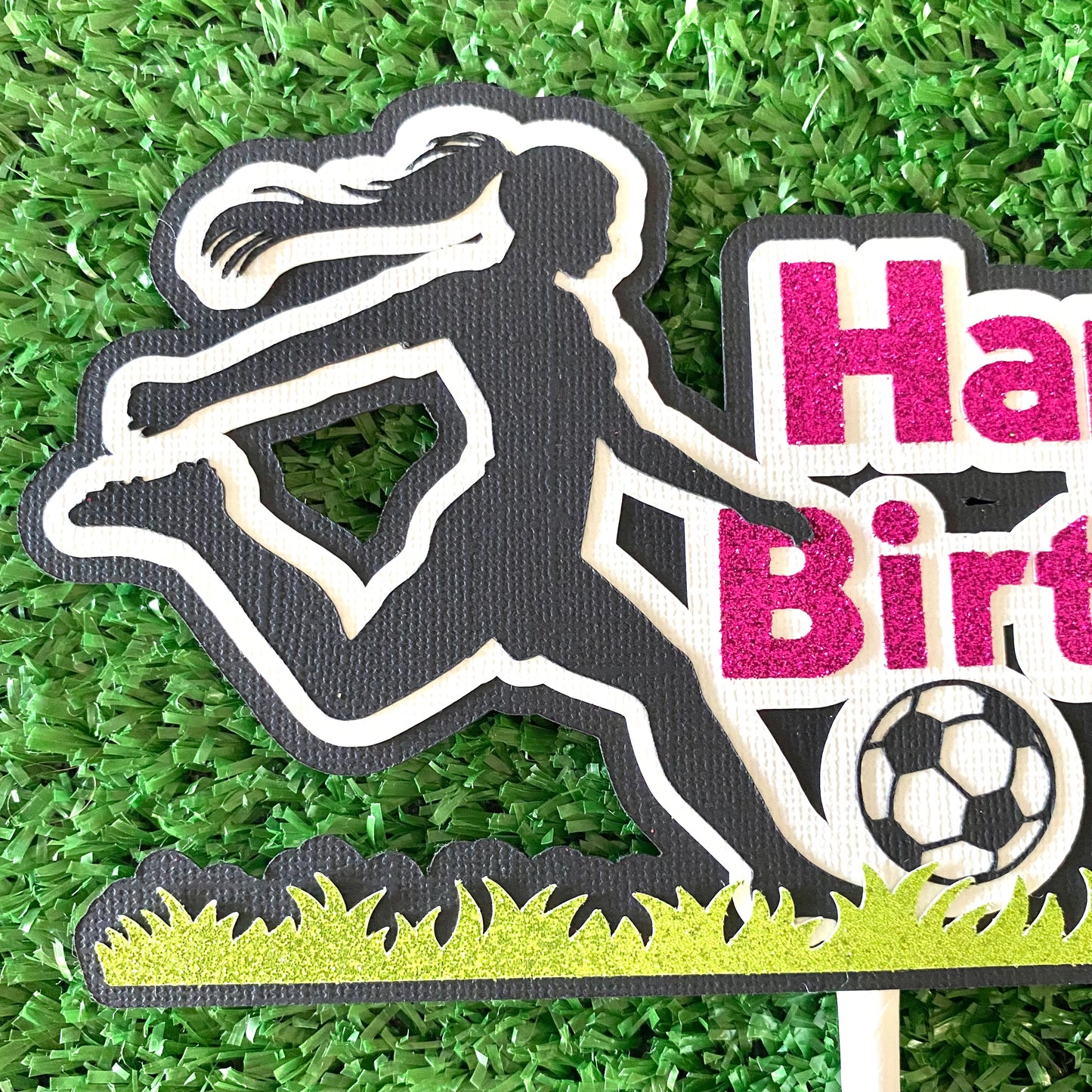 Female cake topper with pink words saying happy birthday and a girl soccer player in black silhouette kicking a soccer ball on glitter green grass.