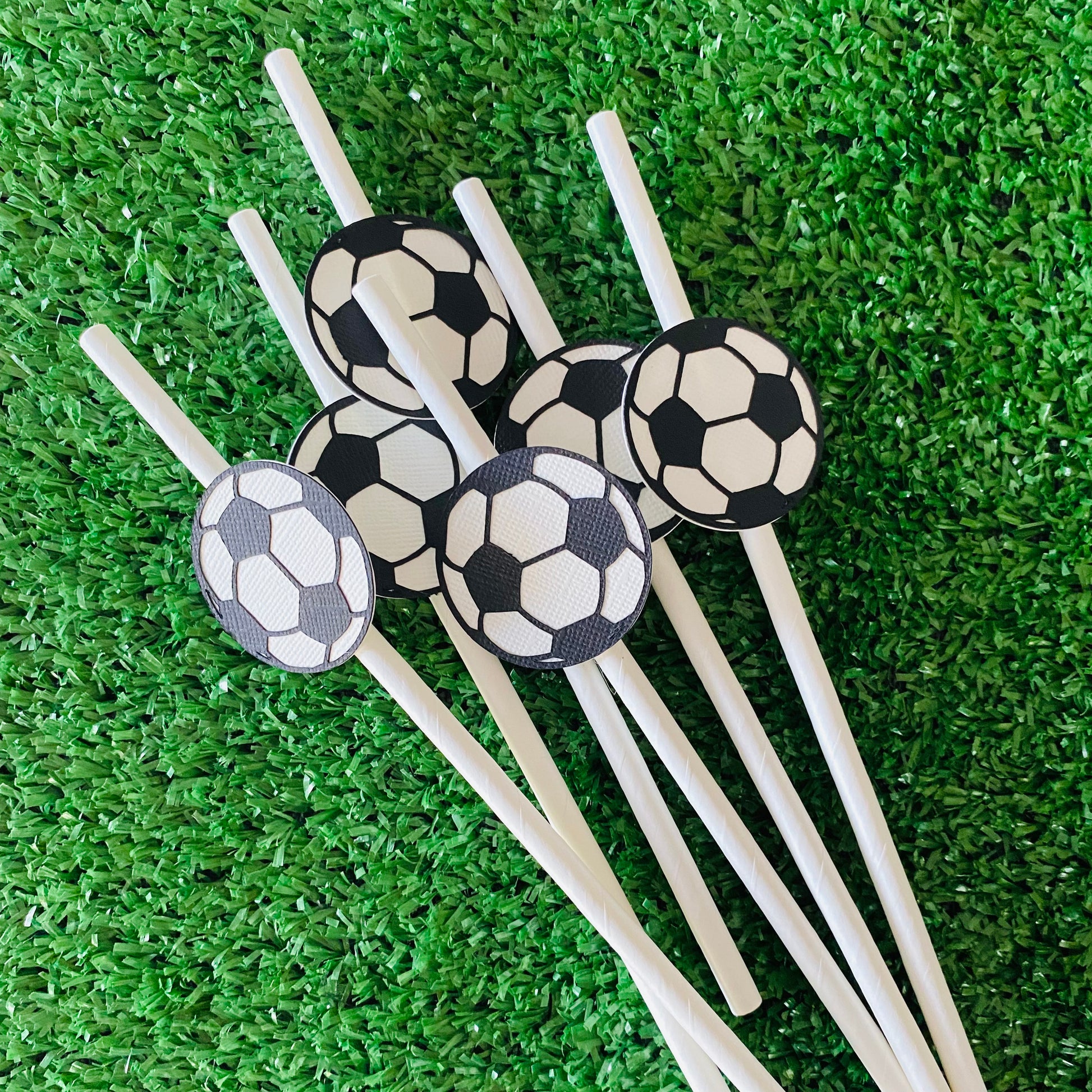 Bunch of white straws topped with soccer ball paper straw toppers laying on grass.