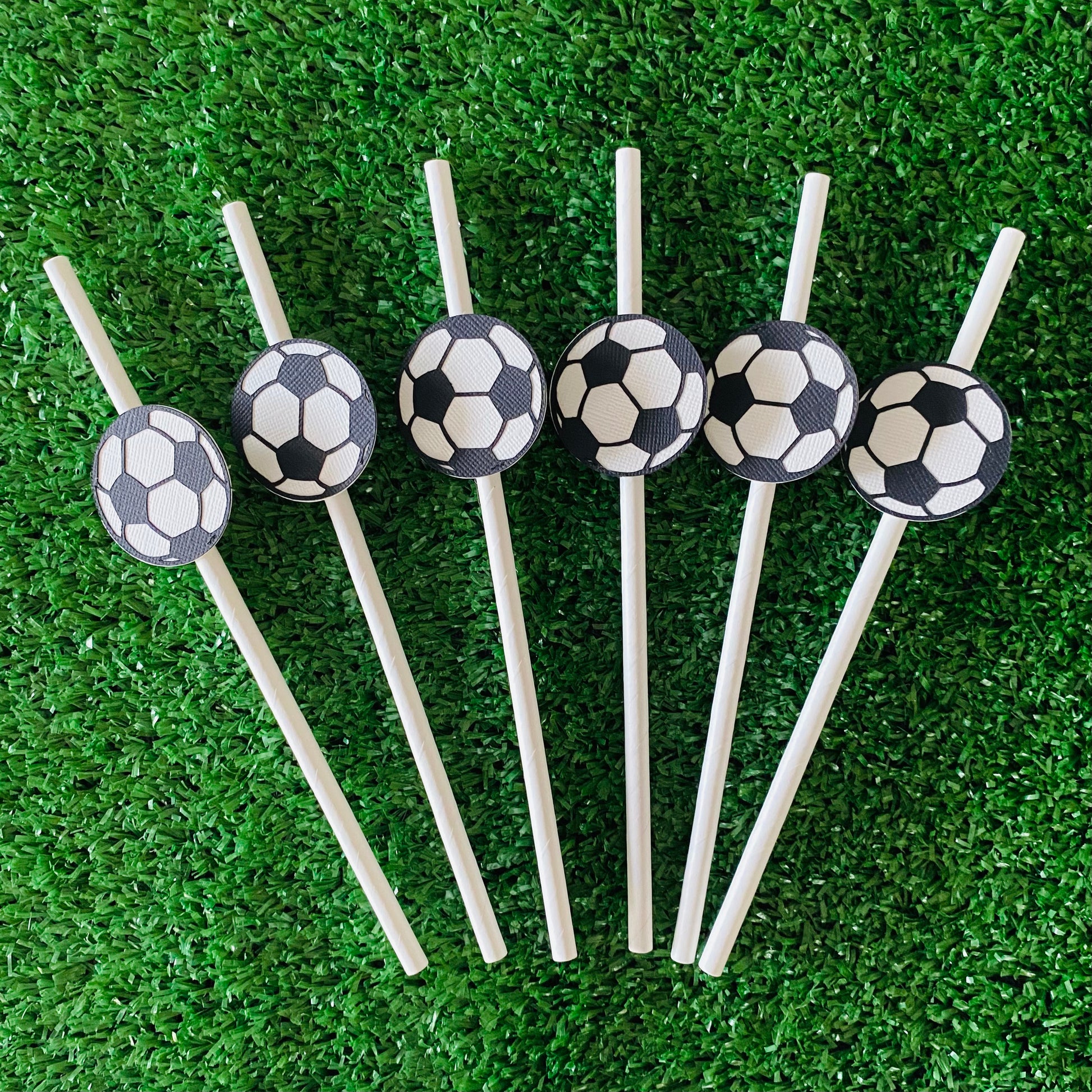 White straws topped with soccer ball paper straw toppers laying on green grass in a fan shape.