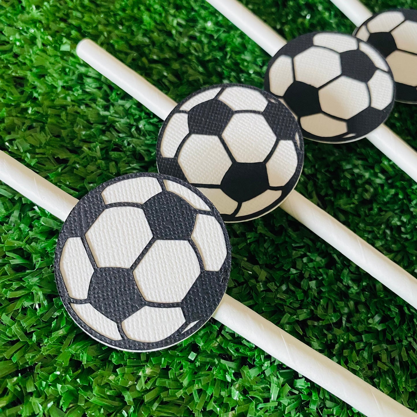 Bunch of white straws topped with soccer ball paper straw toppers laying on grass.