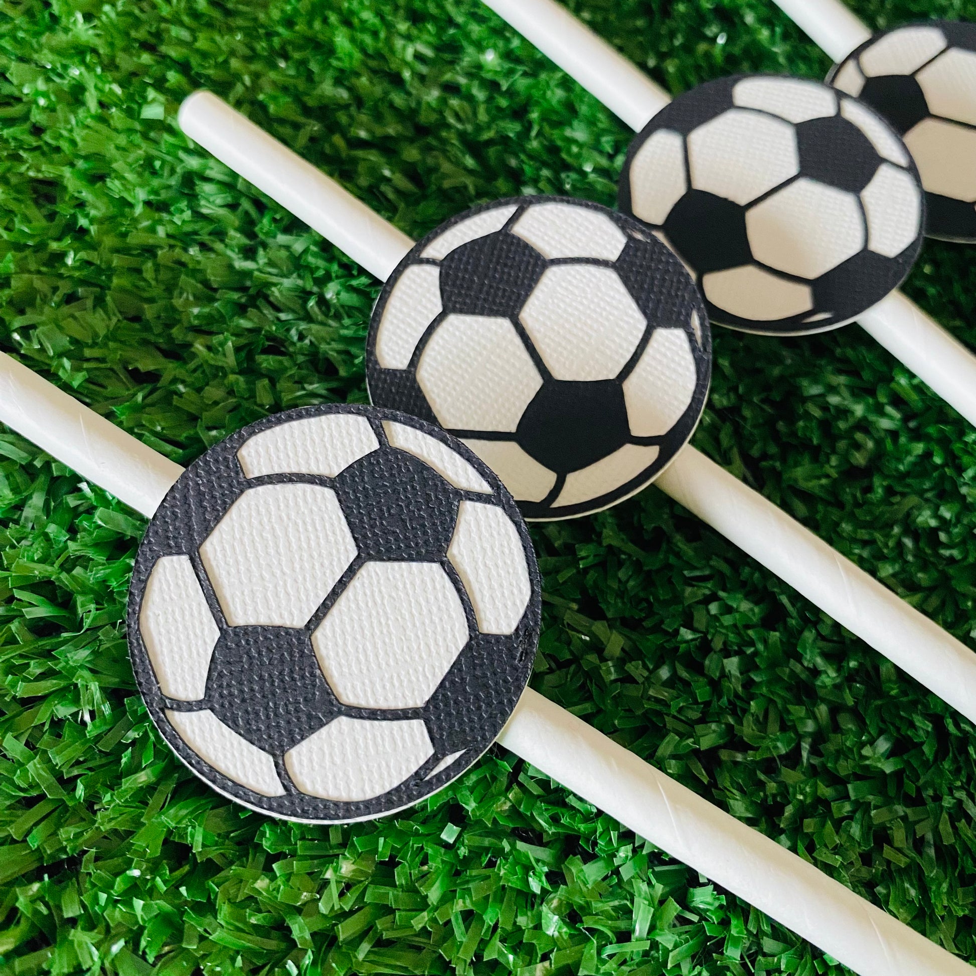 Bunch of white straws topped with soccer ball paper straw toppers laying on grass.
