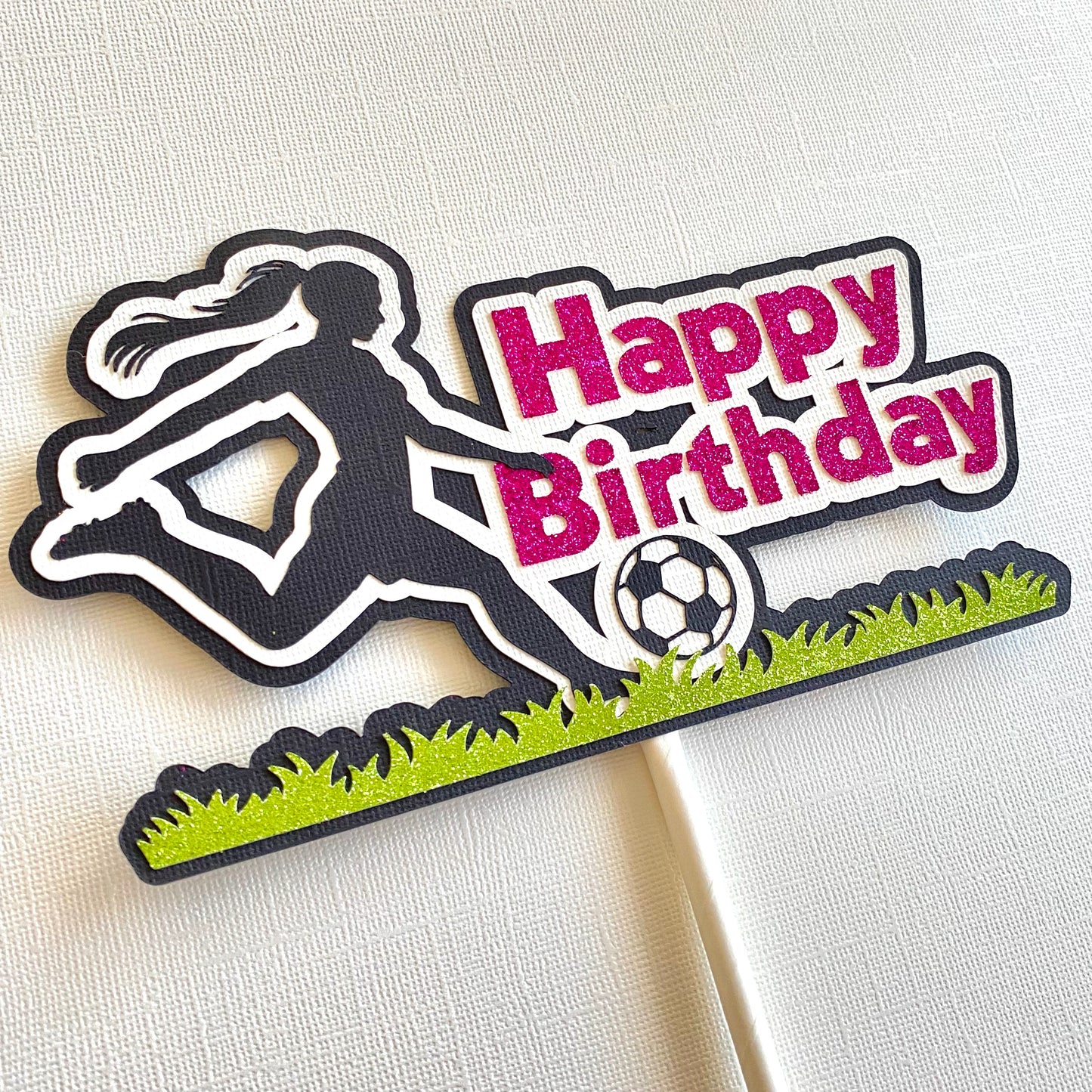 Female cake topper with pink words saying happy birthday and a girl soccer player in black silhouette kicking a soccer ball on glitter green grass laying on a white table.