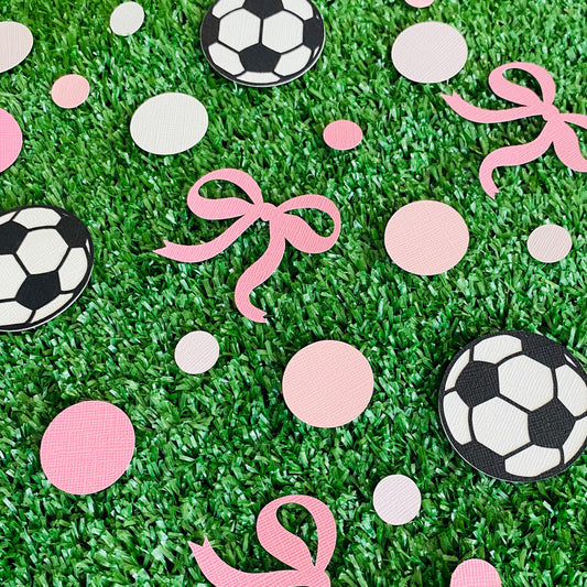 green grass with party scatters of pink and white mixed size circles and black and white soccer balls with added pink bows.