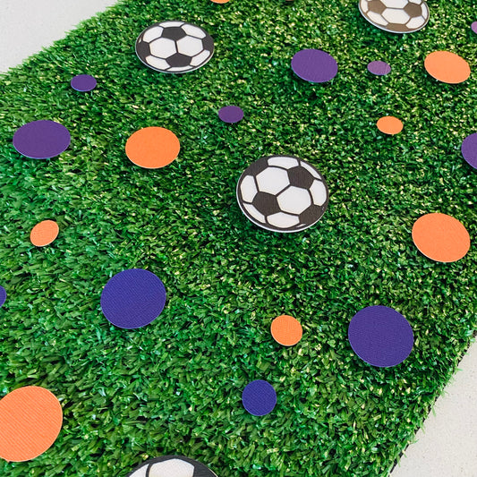 Blue and orange round paper cutouts with soccer ball cutouts spread on synthetic grass