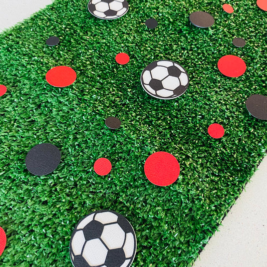 Red and black paper round discs with soccer ball paper discs  scattered across green grass.